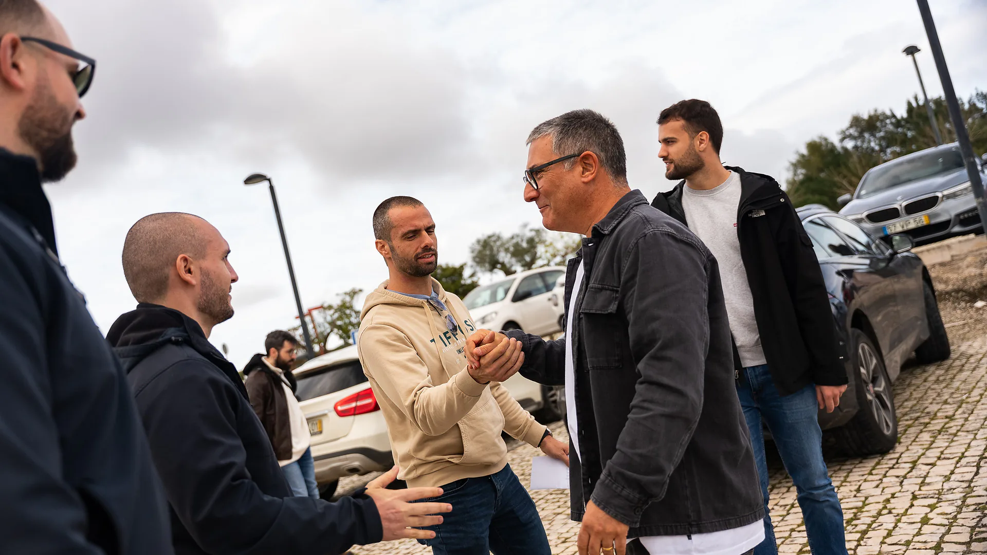 Group of men greeting each other outdoors in a parking area, shaking hands and chatting beside parked cars under a cloudy sky.
