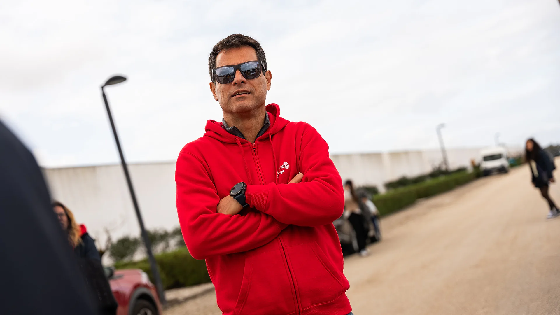 Group of men greeting each other outdoors in a parking area, shaking hands and chatting beside parked cars under a cloudy sky.