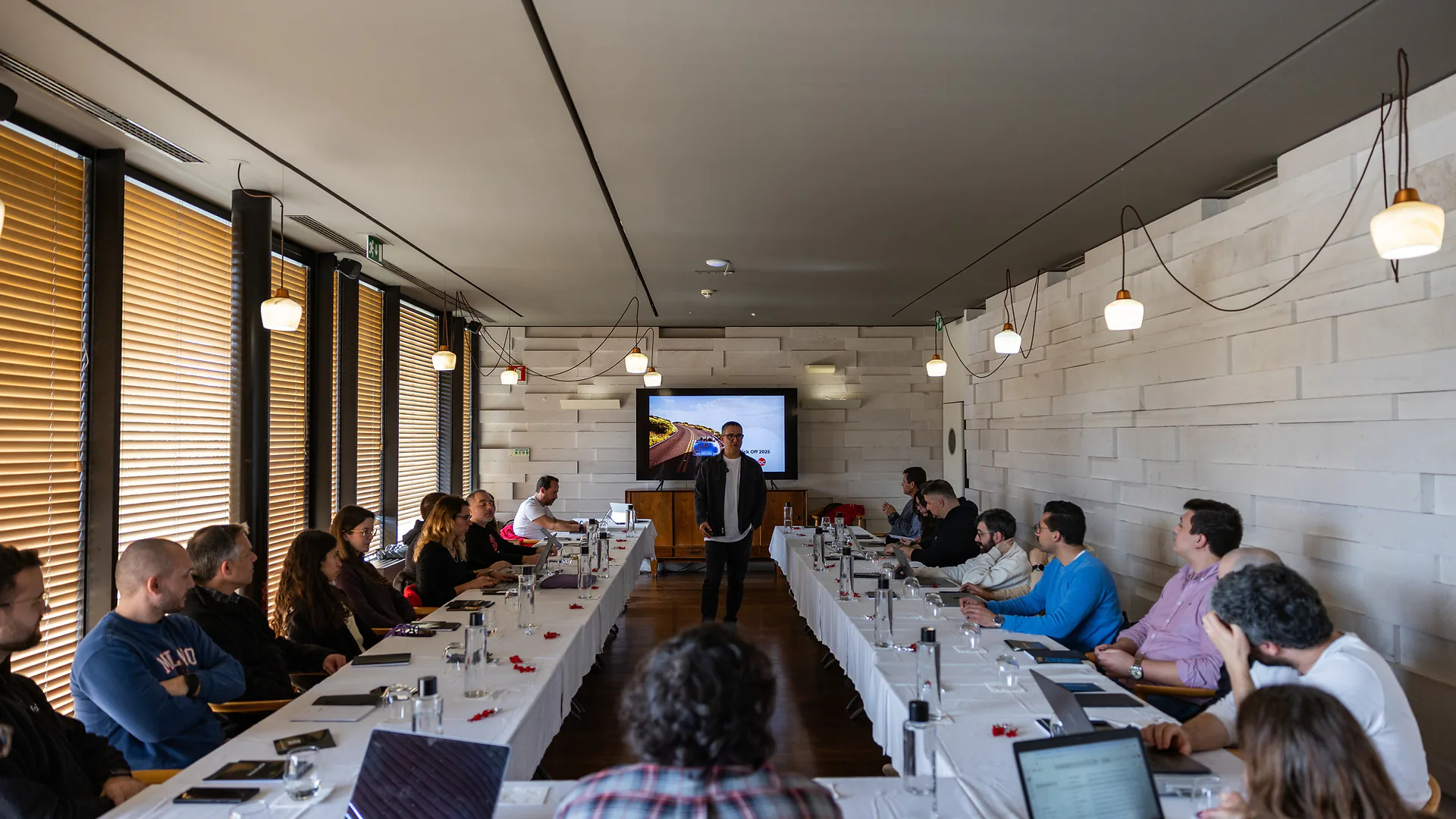 Three men smiling and talking indoors in a dining room set with long tables and chairs, with a large screen displaying a fireplace in the background.