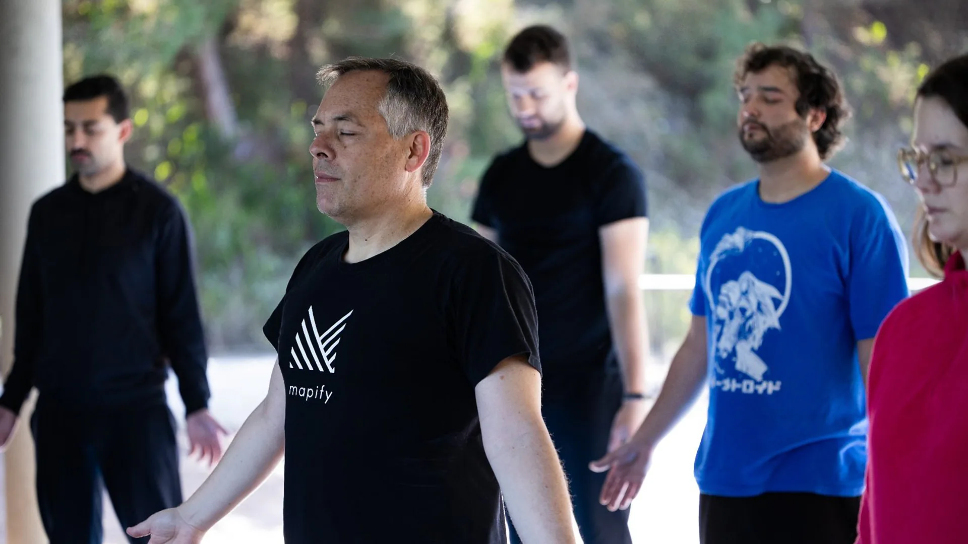 Two men standing at the front of the room during a presentation, one speaking while the other listens beside a laptop on a table.