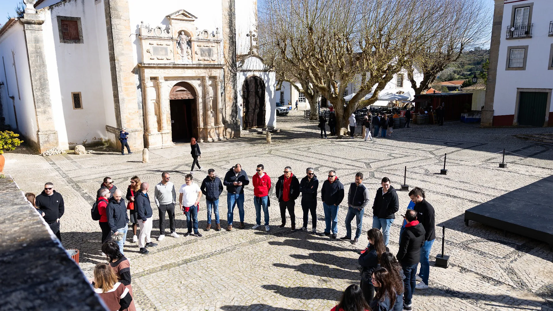 Group of colleagues standing indoors chatting and holding drinks during a karaoke night, with warm lighting and a relaxed atmosphere.
