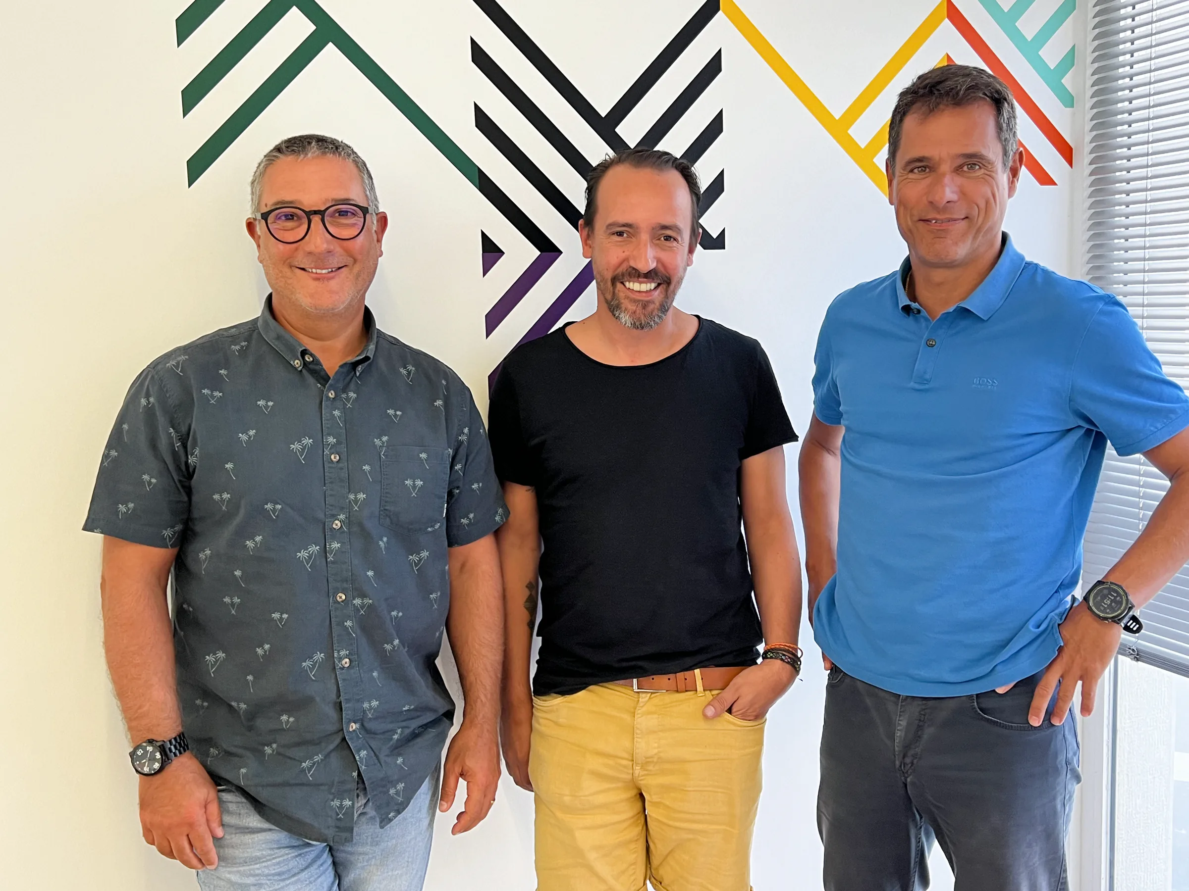 Three men standing side by side in front of a white wall with colorful geometric line graphics, smiling at the camera; one wears glasses and a patterned shirt, the middle wears a black t-shirt with yellow shorts, and the third wears a blue polo shirt.