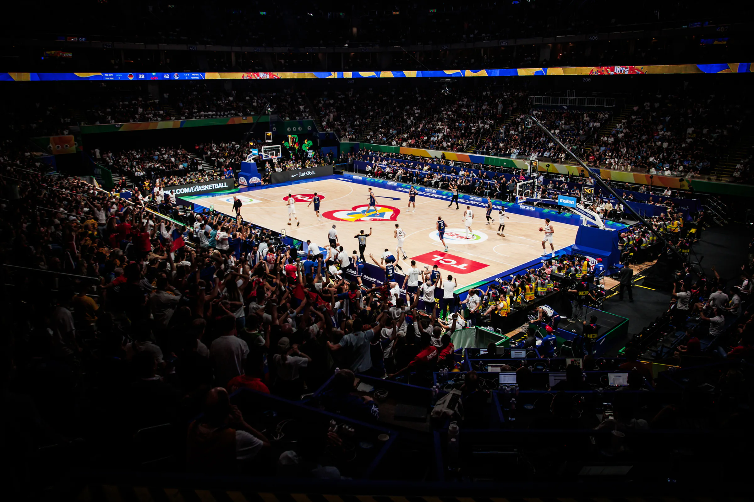 Crowded basketball stadium with teams playing on court and a large cheering crowd in the stands.